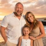 Alex Jones posing with his wife Erika Wulff Jones and their young daughter on a beach during a beautiful sunset.