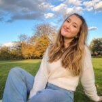A young woman with long wavy blonde hair wearing a white knit sweater and jeans, sitting on the grass in a park on a sunny day.
