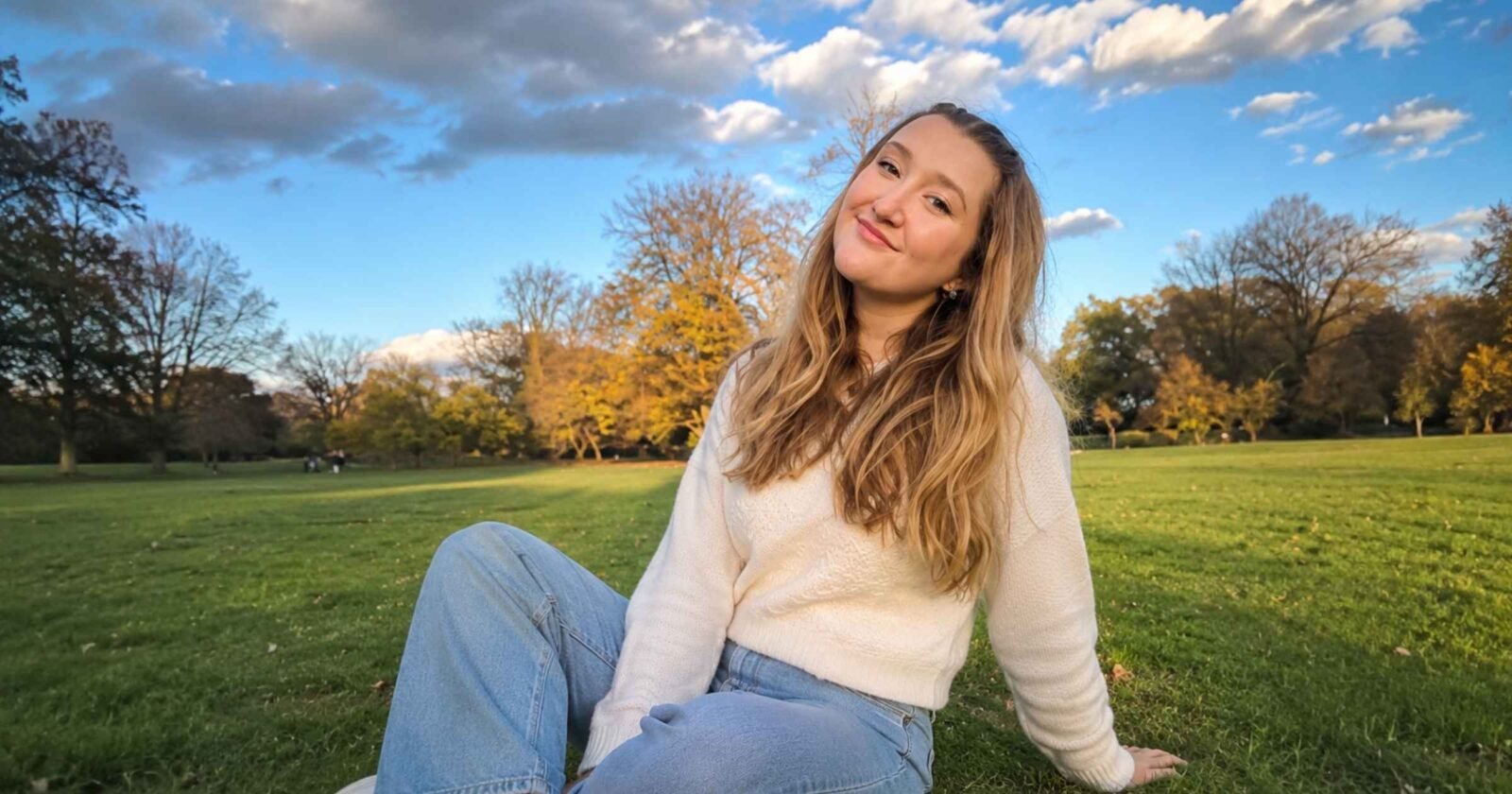 A young woman with long wavy blonde hair wearing a white knit sweater and jeans, sitting on the grass in a park on a sunny day.
