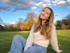A young woman with long wavy blonde hair wearing a white knit sweater and jeans, sitting on the grass in a park on a sunny day.