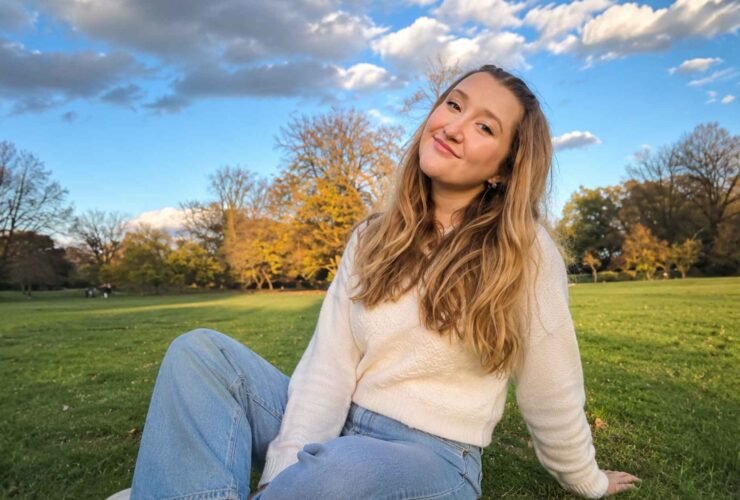 A young woman with long wavy blonde hair wearing a white knit sweater and jeans, sitting on the grass in a park on a sunny day.