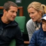 A young Huckleberry Grylls wearing a blue puffer jacket and white cap sitting between his parents, Bear and Shara Grylls, at a sporting event.