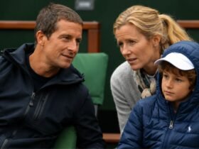 A young Huckleberry Grylls wearing a blue puffer jacket and white cap sitting between his parents, Bear and Shara Grylls, at a sporting event.