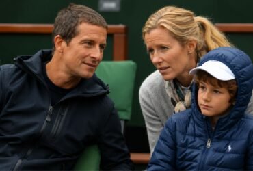 A young Huckleberry Grylls wearing a blue puffer jacket and white cap sitting between his parents, Bear and Shara Grylls, at a sporting event.