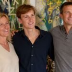 Jesse Grylls smiling and standing confidently between his parents, Shara and Bear Grylls, in front of a colorful abstract expressionist painting.