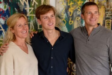 Jesse Grylls smiling and standing confidently between his parents, Shara and Bear Grylls, in front of a colorful abstract expressionist painting.