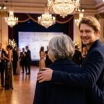 Actor Joe Alwyn smiling gently with his arm around his mother, Elizabeth Alwyn, who is viewed from the back at a formal gala event hall.
