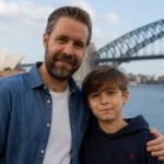 Actor Paddy Considine standing next to his son, musician Joseph Considine, smiling together with the Sydney Harbour Bridge in the background.