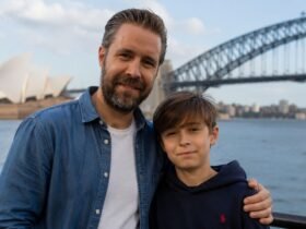 Actor Paddy Considine standing next to his son, musician Joseph Considine, smiling together with the Sydney Harbour Bridge in the background.