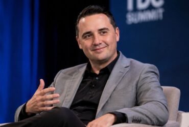 A professional portrait of a smiling male attorney wearing a grey suit jacket and black collared shirt during a speaking summit.