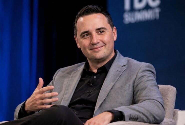 A professional portrait of a smiling male attorney wearing a grey suit jacket and black collared shirt during a speaking summit.