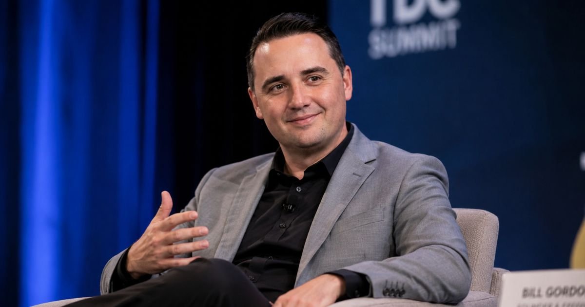 A professional portrait of a smiling male attorney wearing a grey suit jacket and black collared shirt during a speaking summit.