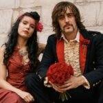 Rian Haynes, A vintage-style portrait of a bride in a red dress and a groom in a ruffled shirt and dark jacket holding a bouquet of red roses.