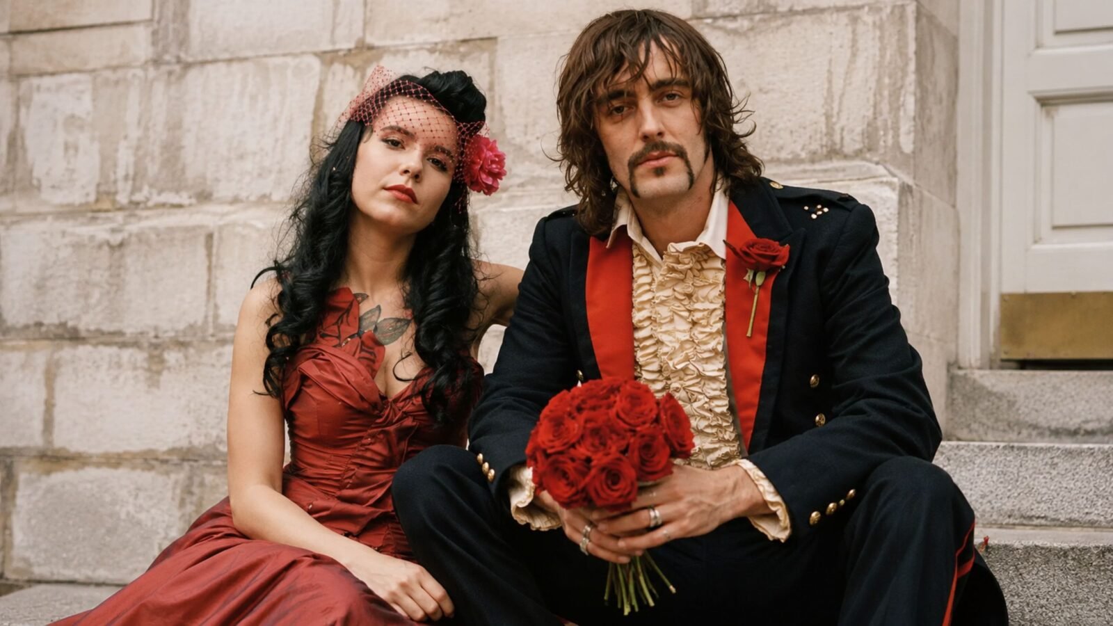 Rian Haynes, A vintage-style portrait of a bride in a red dress and a groom in a ruffled shirt and dark jacket holding a bouquet of red roses.