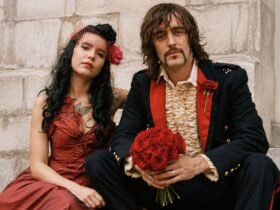 Rian Haynes, A vintage-style portrait of a bride in a red dress and a groom in a ruffled shirt and dark jacket holding a bouquet of red roses.