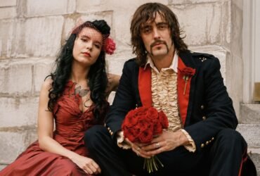 Rian Haynes, A vintage-style portrait of a bride in a red dress and a groom in a ruffled shirt and dark jacket holding a bouquet of red roses.
