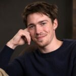 Portrait of emerging British actor Patrick Alwyn smiling gently while resting his head on his hand.