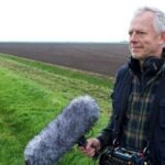 Documentary filmmaker Richard Alwyn holding a professional boom microphone and wearing headphones while filming in an open field.