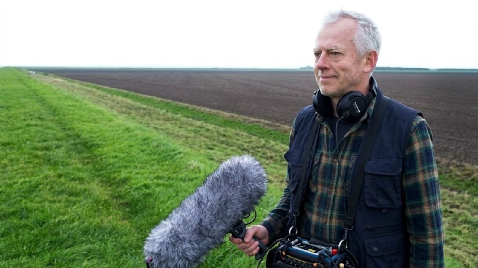 Documentary filmmaker Richard Alwyn holding a professional boom microphone and wearing headphones while filming in an open field.