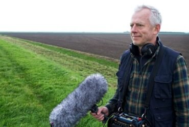 Documentary filmmaker Richard Alwyn holding a professional boom microphone and wearing headphones while filming in an open field.